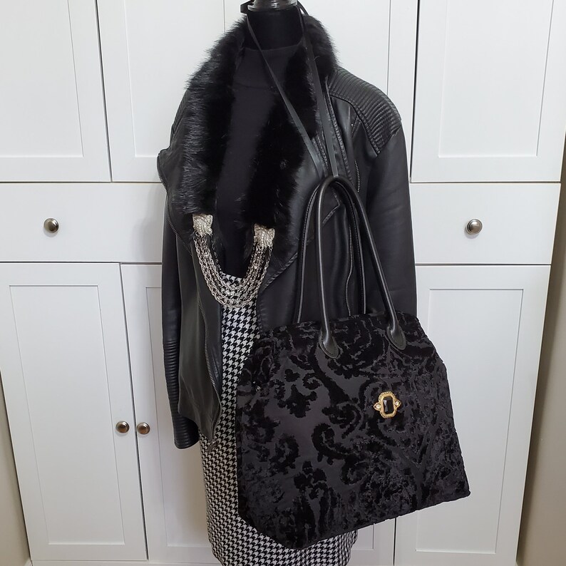 Image of black jacquard velvet carpet bag displayed on a mannequin. Bag is created with upholstery fabric. The bag features top handles in black leather. Bag is decorated with a vintage gold tone brooch framing a sparkling black faux gemstone.