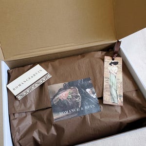 May include: An open cardboard shipping box containing brown tissue paper, a rectangular white card with the text ROMANCE & RUIN, a square card featuring a floral image and the text ROMANCE & RUIN, and a vertical bookmark with a vintage illustration.