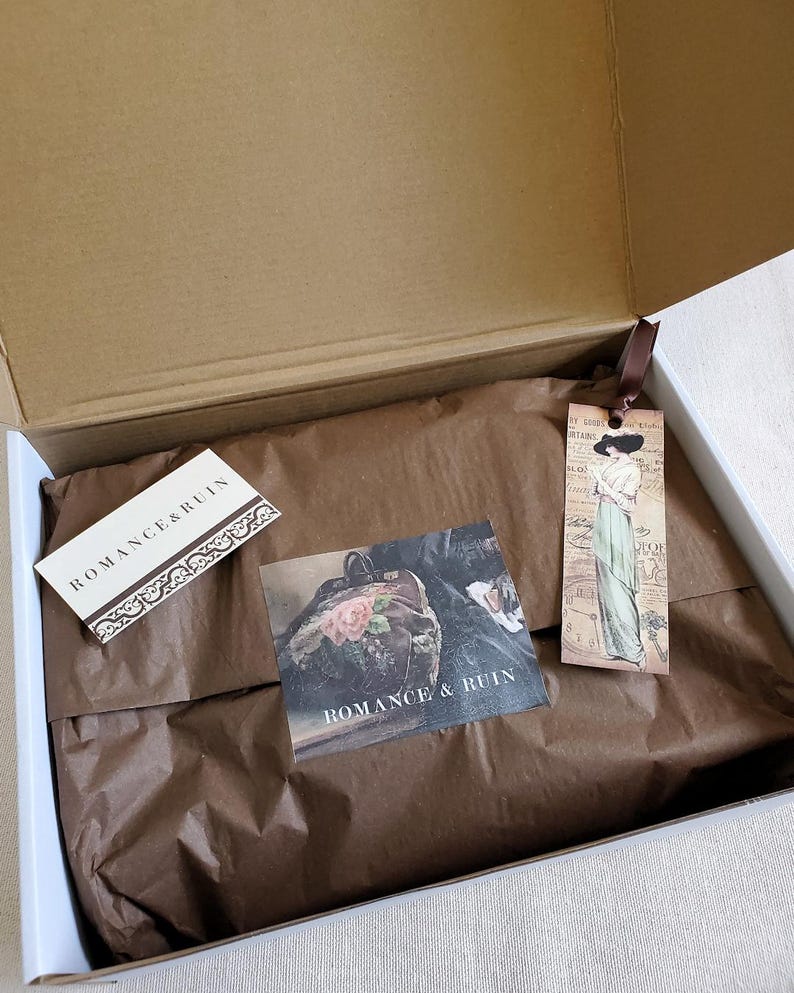 May include: An open cardboard shipping box containing brown tissue paper, a rectangular white card with the text ROMANCE & RUIN, a square card featuring a floral image and the text ROMANCE & RUIN, and a vertical bookmark with a vintage illustration.