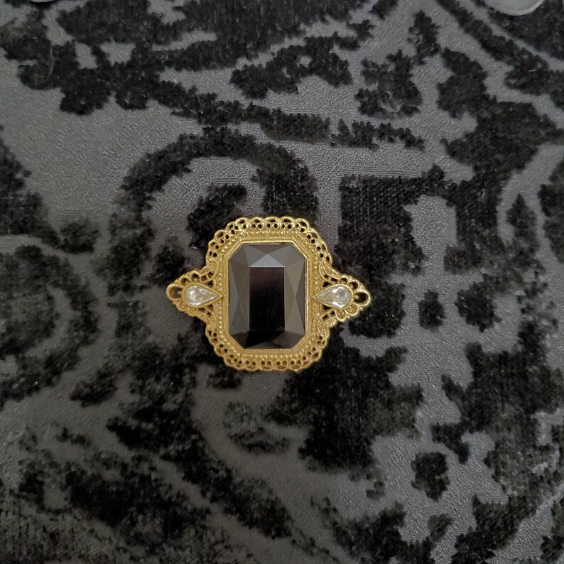 May include: A gold-toned brooch featuring a large, rectangular, faceted black center stone surrounded by an intricate, filigree-style metal frame. Two smaller, clear, pear-shaped stones are set on the left and right sides of the central piece.