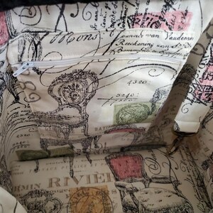 May include: The interior of a fabric bag featuring a cream-colored lining with a black and pink print of vintage chairs, cursive French script, and postage stamp motifs. It includes an integrated zippered pocket with a white zipper pull.