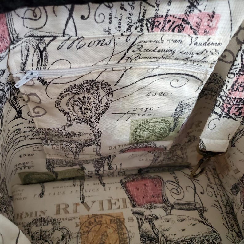 May include: The interior of a fabric bag featuring a cream-colored lining with a black and pink print of vintage chairs, cursive French script, and postage stamp motifs. It includes an integrated zippered pocket with a white zipper pull.