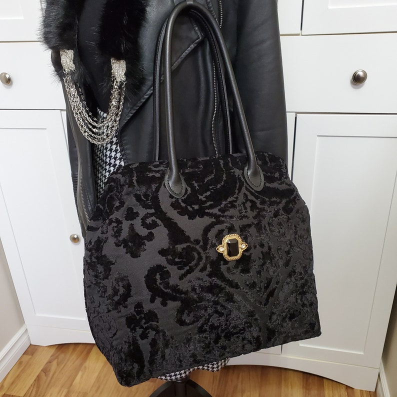 Image of black jacquard velvet carpet bag displayed on a mannequin. Bag is created with upholstery fabric. The bag features top handles in black leather. Bag is decorated with a vintage gold tone brooch framing a sparkling black faux gemstone.