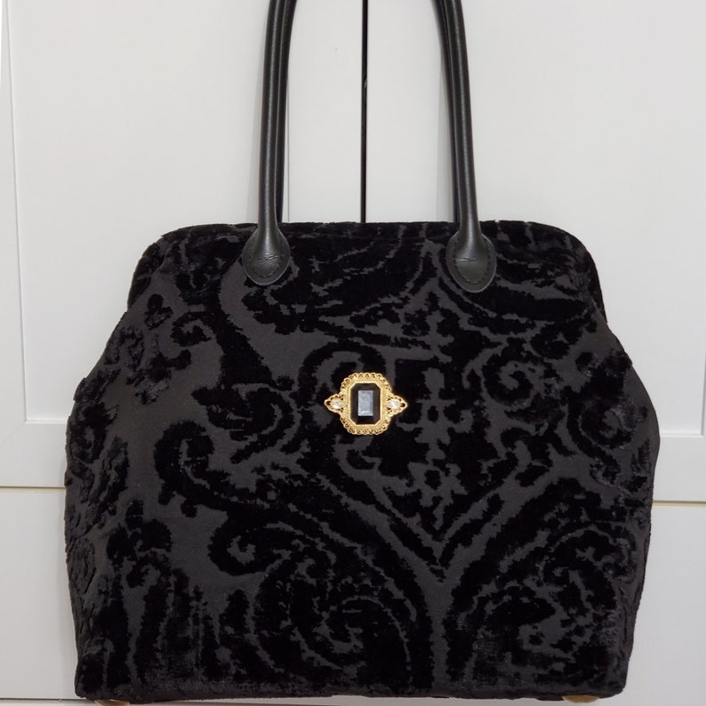 Close up image of black jacquard velvet carpet bag created with upholstery fabric in a damask pattern. The bag features top handles in black leather. bag is decorated with a vintage gold tone brooch framing a sparkling black faux gemstone.
