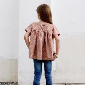 May include: A young girl wearing a brown and white polka dot blouse with short sleeves and blue jeans. She is standing in front of a white wall.