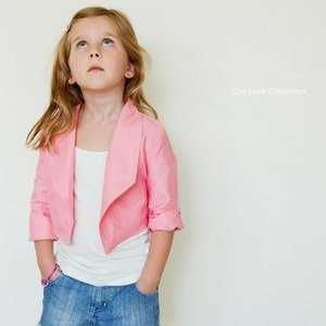 以下が含まれることがあります： ピンクのクロップドジャケットに白いシャツと青いジーンズを着用した若い女の子。ジャケットはドレープされた襟と長袖です。女の子は上を見上げて微笑んでいます。