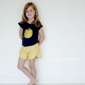 May include: A young girl wearing a navy blue t-shirt with a yellow apple graphic and yellow shorts with a white floral pattern. The text "CALI FAYE COLLECTION." is visible in the bottom right corner of the image.