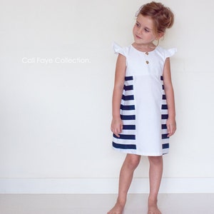 May include: A young girl wearing a white dress with a black and white striped panel. The dress has a round neckline and short sleeves with ruffles. The text "Cali Faye Collection." is visible in the top left corner of the image.