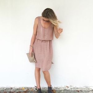 May include: A woman wearing a blush pink sleeveless dress with a drawstring waist and a gold necklace. She is wearing gray ankle boots and holding a gold clutch.
