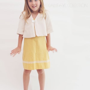 Può includere: Una bambina indossa un cardigan bianco sopra un vestito giallo a pois con un bordo di pizzo bianco. Indossa scarpe leopardate con accenti rosa. Il testo "Cali Faye Collection" è visibile sullo sfondo.