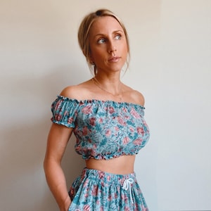 May include: A teal two-piece set, comprising an off-the-shoulder crop top and a skirt, both with a floral design in pink and red. The skirt has a drawstring waist. The outfit is a coordinated ensemble.
