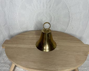 May include: A small, brass-colored bell with a loop handle sits on a round, light-colored wooden stand. The bell has a classic shape, and the stand has three small legs. The background is a white, textured fabric.