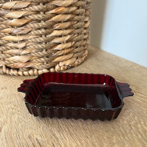 Könnte beinhalten: Eine kleine, rechteckige, dunkelrote Glasschale mit geriffelten Rändern und zwei kleinen Griffen an jeder Seite.