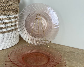 Puede incluir: Dos platos de cristal rosa con diseño en espiral, uno expuesto en un soporte dorado. Los platos tienen un motivo floral en el centro. Se ve una cesta tejida en el fondo.