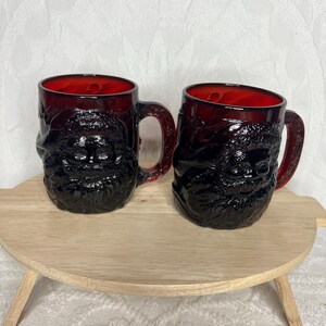 Op de afbeelding: Twee donkerrode glazen mokken met een gedetailleerd gezichtsontwerp. De mokken hebben een handvat en een rode binnenkant. Ze worden gepresenteerd op een licht houten dienblad.