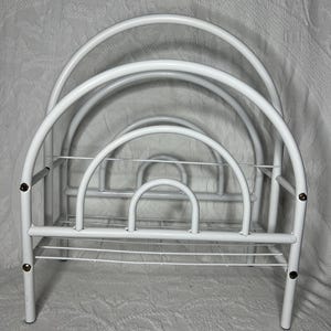 May include: A white metal magazine rack with a series of arched supports. The rack has a rectangular base and a wire grid at the bottom. The design is simple and functional, suitable for organizing reading materials.