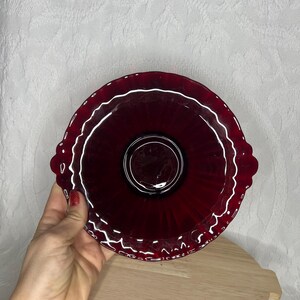 Può includere: Un piatto da portata in vetro rosso rubino con bordo smerlato e due piccole maniglie. Il piatto ha un design circolare con un centro rialzato ed è appoggiato su un vassoio di legno.