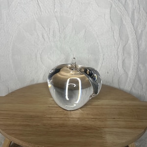 May include: A clear glass apple sculpture with a stem and a leaf-like detail. The apple is sitting on a small, round wooden table. The background is a white, textured fabric.