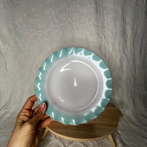 Può includere: Un piatto in ceramica bianca con un bordo smerlato azzurro. Il piatto è tenuto in mano e poggia su un piccolo supporto rotondo in legno. Lo sfondo è un tessuto bianco strutturato.