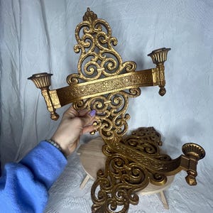Puede incluir: Aplique de pared dorado y ornamentado con dos candelabros. El aplique presenta intrincados diseños de volutas y florales. Los candelabros están colocados a ambos lados de una barra horizontal. El diseño general es simétrico y decorativo.