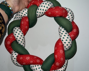 May include: Braided Christmas wreath with red fabric featuring white snowflakes, green fabric, and white fabric with green Christmas trees. The wreath is held by a hand against a white background. A red string is attached for hanging.