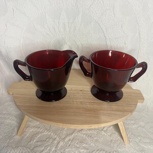 May include: A ruby red glass creamer and sugar bowl set. The creamer has a spout and handle, while the sugar bowl has a handle on each side. Both pieces sit on a small, round wooden tray with legs. The set is displayed against a white background.