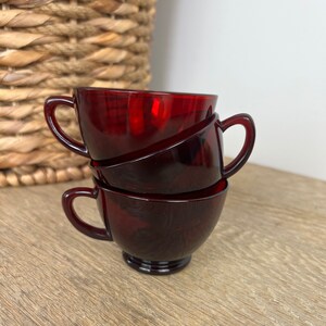 Könnte beinhalten: Drei rote Glasbecher, die übereinander gestapelt sind. Die Becher haben eine abgerundete Form und einen Henkel an jeder Seite.