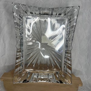 May include: A clear, rectangular glass picture frame with a decorative, sunburst-like design in the center. The frame has a ridged border and is set on a small, round wooden table. The frame is designed to hold a photograph.