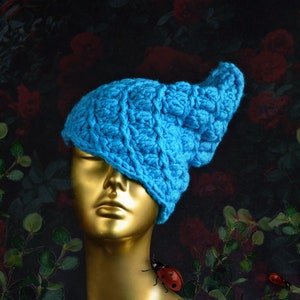 Op de afbeelding: Een fel turquoise blauwe muts met een gestructureerd, verhoogd bobbelsteekpatroon. De muts wordt getoond op een metallic goudkleurig etalagehoofd, dat een glad, reflecterend oppervlak en neutrale gelaatstrekken heeft.