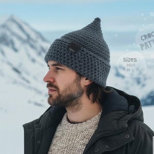 May include: A gray crochet hat with a pointed top, worn by a person. The hat has a small, dark patch on the front. The image includes text that says "CROCHET PATTERN" and "SIZES ADULT S M L XL". The background shows a snowy mountain.