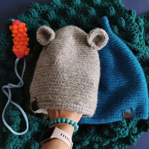 May include: A gray knit hat with attached ears and a blue knit hat are displayed on a textured green blanket. A small orange and pink carrot toy with a blue string is also present. The hats have a tag that says "Ulou".