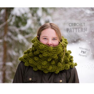 May include: A green crocheted cowl with a textured, bobble stitch design. The cowl is worn around the neck and shoulders. The image also includes the text "CROCHET PATTERN" and "Printable PDF 9 pages".