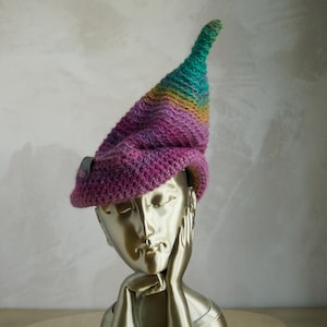 Può includere: Un cappello all'uncinetto con una parte superiore a punta e una tesa larga, caratterizzato da un motivo a colori sfumati che passa dal viola alla base al rosa, giallo e verde sulla punta. Una piccola etichetta rettangolare grigia è fissata sul lato.
