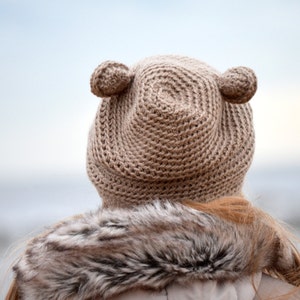 May include: A tan crocheted hat with two small, round ear-like accents on top. The hat is worn by a person with long, light brown hair and a fur-trimmed coat. The background is a blurred view of the ocean.