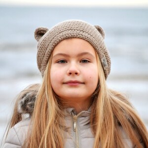 May include: A young person with long blonde hair wearing a tan knit hat with bear ears. The person is also wearing a light-colored jacket with a fur-lined hood. The background is a blurred view of the ocean.