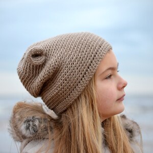 May include: A tan crocheted beanie hat with a unique swirl detail. The hat is made of a textured yarn and is worn by a person with long blonde hair. The background is a blurred view of the sky and water.