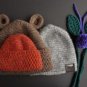 May include: Three knitted hats in brown, orange, and gray. The brown hat has bear ears. The orange hat is nested inside the brown hat. The gray hat has a small leather tag. A green and purple knitted decoration is on the right.