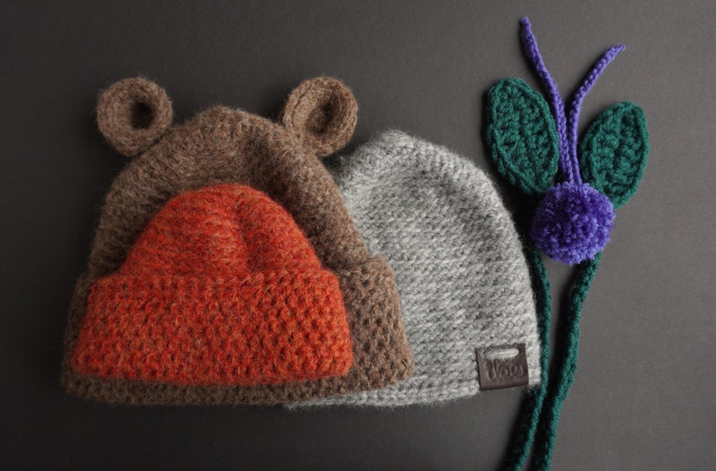 May include: Three knitted hats in brown, orange, and gray. The brown hat has bear ears. The orange hat is nested inside the brown hat. The gray hat has a small leather tag. A green and purple knitted decoration is on the right.
