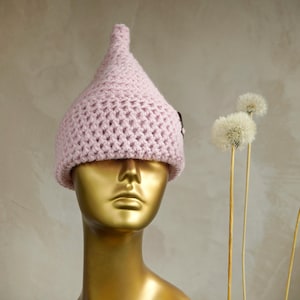 May include: A light pink crocheted hat with a pointed top and a textured, woven stitch pattern. The hat is displayed on a smooth, metallic gold-coloured mannequin head, showcasing the soft yarn construction and the distinct conical shape.