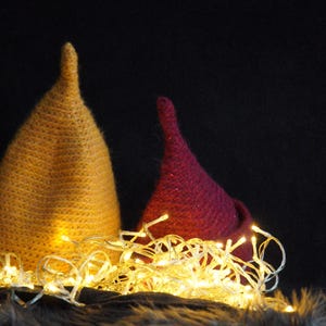 Op de afbeelding: Twee puntige, kegelvormige mutsen, &eacute;&eacute;n in mosterdgeel en &eacute;&eacute;n in donker magenta, zijn geplaatst achter een cluster van gloeiende, warmwitte lichtsnoeren. De mutsen hebben een getextureerd, gebreid uiterlijk met een duidelijke, taps toelopende punt.