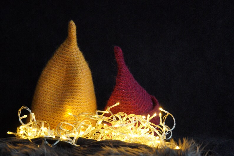 Op de afbeelding: Twee puntige, kegelvormige mutsen, &eacute;&eacute;n in mosterdgeel en &eacute;&eacute;n in donker magenta, zijn geplaatst achter een cluster van gloeiende, warmwitte lichtsnoeren. De mutsen hebben een getextureerd, gebreid uiterlijk met een duidelijke, taps toelopende punt.