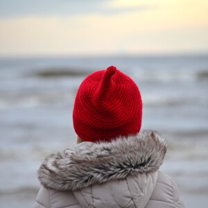 A person wearing a red knitted hat with a pointed top, standing in front of a body of water. The hat is a bright red colour and has a fuzzy texture. The person is wearing a grey jacket with a fur collar.