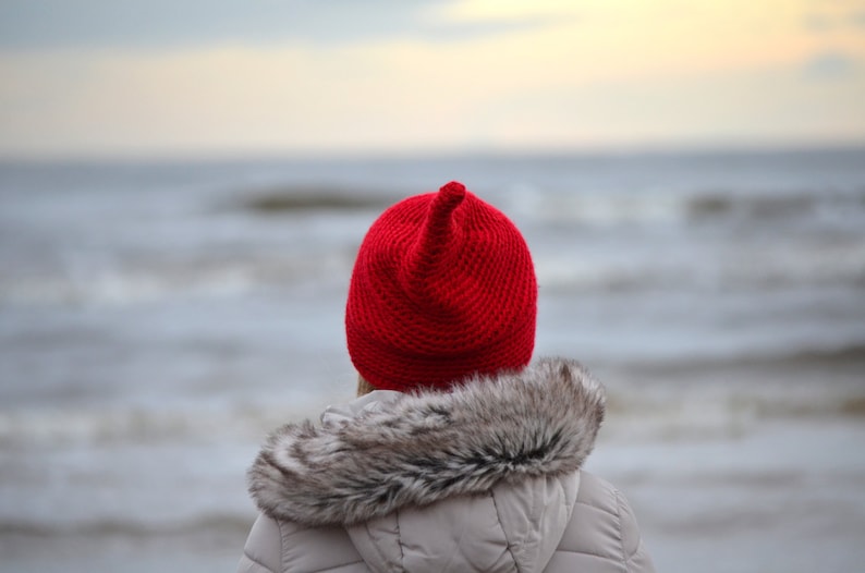 A person wearing a red knitted hat with a pointed top, standing in front of a body of water. The hat is a bright red colour and has a fuzzy texture. The person is wearing a grey jacket with a fur collar.