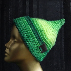 May include: A green and lime green crocheted hat with a pointed top. The hat features a gradient of green shades and a small brown leather tag. The hat is displayed on a mannequin head against a dark background.