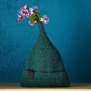 Op de afbeelding: Een donkergroene gebreide muts met een hoge, puntige kroon en een omgeslagen rand. Een klein bruin leren label met de tekst "Vlas" is aan de voorzijde bevestigd. Een takje met lichtpaarse seringenbloemen steekt boven in de puntige muts.