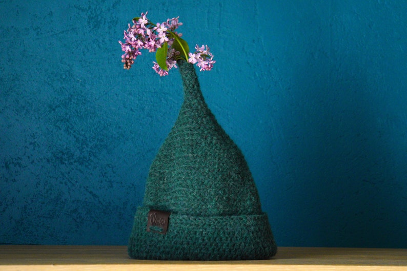 Op de afbeelding: Een donkergroene gebreide muts met een hoge, puntige kroon en een omgeslagen rand. Een klein bruin leren label met de tekst "Vlas" is aan de voorzijde bevestigd. Een takje met lichtpaarse seringenbloemen steekt boven in de puntige muts.
