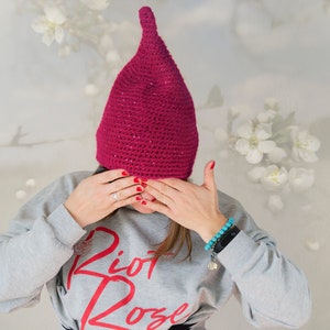 Op de afbeelding: Een magentakleurige gehaakte muts met een puntige bovenkant en subtiele glitteraccenten. De muts wordt gedragen met een lichtgrijze sweater met lange mouwen en de rode tekst "Riot Rose" op de voorzijde, inclusief een kleine rode roosafbeelding.