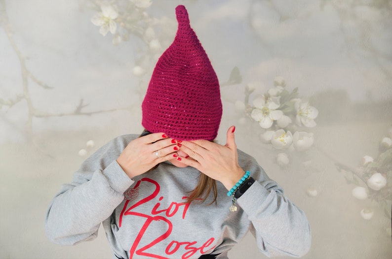 Op de afbeelding: Een magentakleurige gehaakte muts met een puntige bovenkant en subtiele glitteraccenten. De muts wordt gedragen met een lichtgrijze sweater met lange mouwen en de rode tekst "Riot Rose" op de voorzijde, inclusief een kleine rode roosafbeelding.