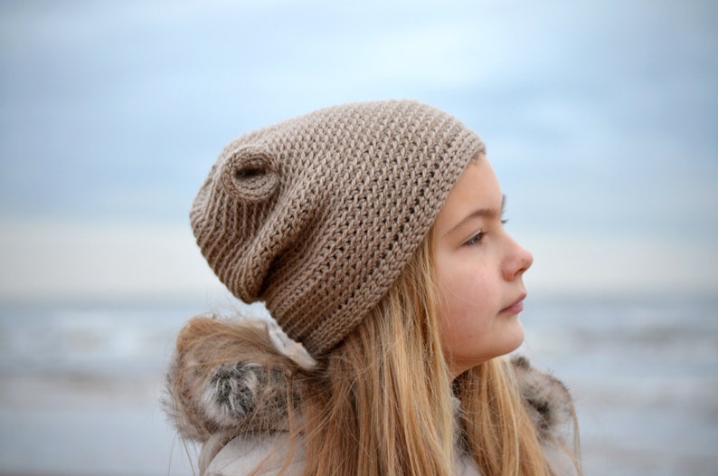 May include: A tan crocheted beanie hat with a unique swirl detail. The hat is made of a textured yarn and is worn by a person with long blonde hair. The background is a blurred view of the sky and water.