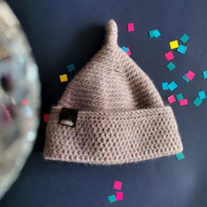 Op de afbeelding: Een licht mauve, getextureerde gebreide muts met een puntige bovenkant en een omgeslagen rand. Een klein, rechthoekig zwart label is bevestigd aan de linkerkant van de rand. De muts heeft een dicht, uniform steekpatroon en een zachte uitstraling.